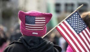 The back of the head of a person carrying an American flag, wearing a pink knitted "pussy hat" with an American flag on it