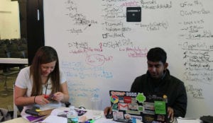 Two hackathon participants sitting at a table in front of a whiteboard with ideas