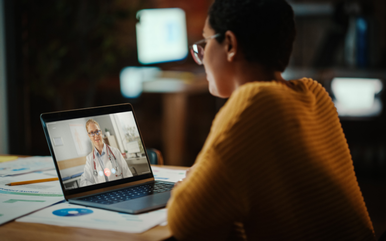 Young woman sitting at table talking with doctor via telehealth appointment on her laptop