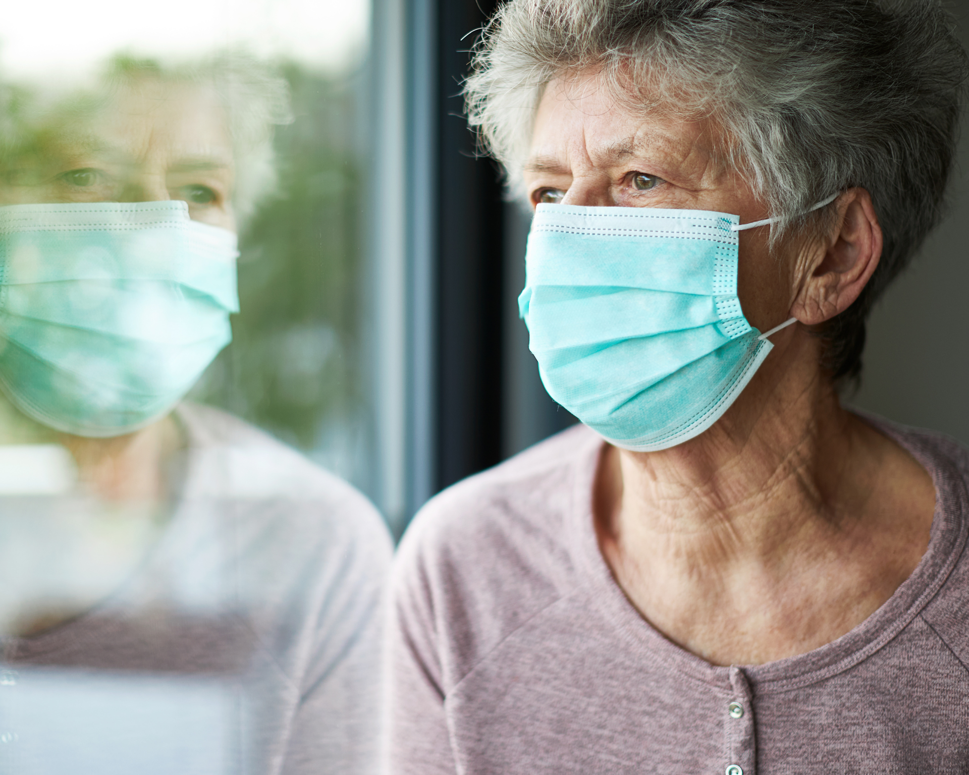 An older woman wearing a surgical mask looks out of the window during the COVID-19 pandemic