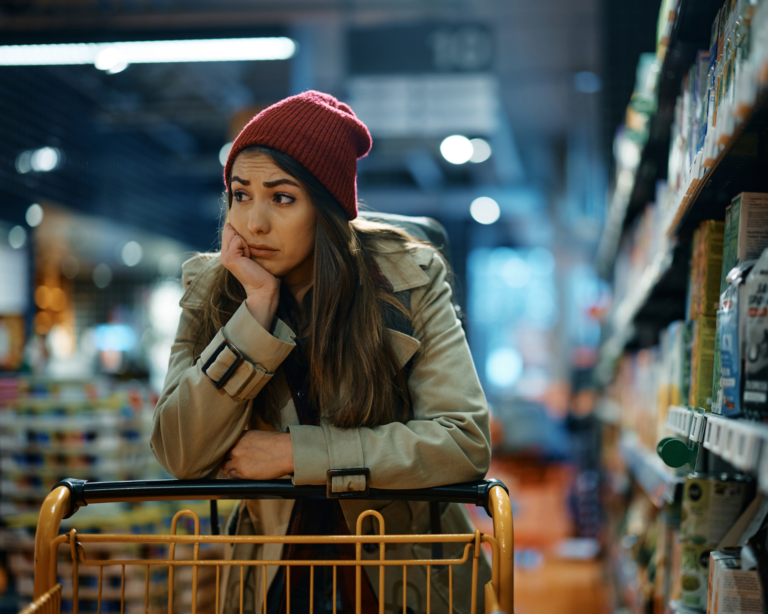 Young sad woman with empty shopping cart among produce aisle at supermarket