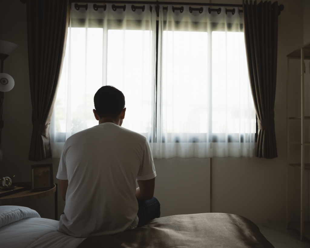 Silhouette of a man sadly sitting on the bed in the bedroom, looking out the window. Depression concept