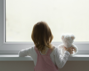 back view of a young child holding a white teddy bear and standing alone at the window