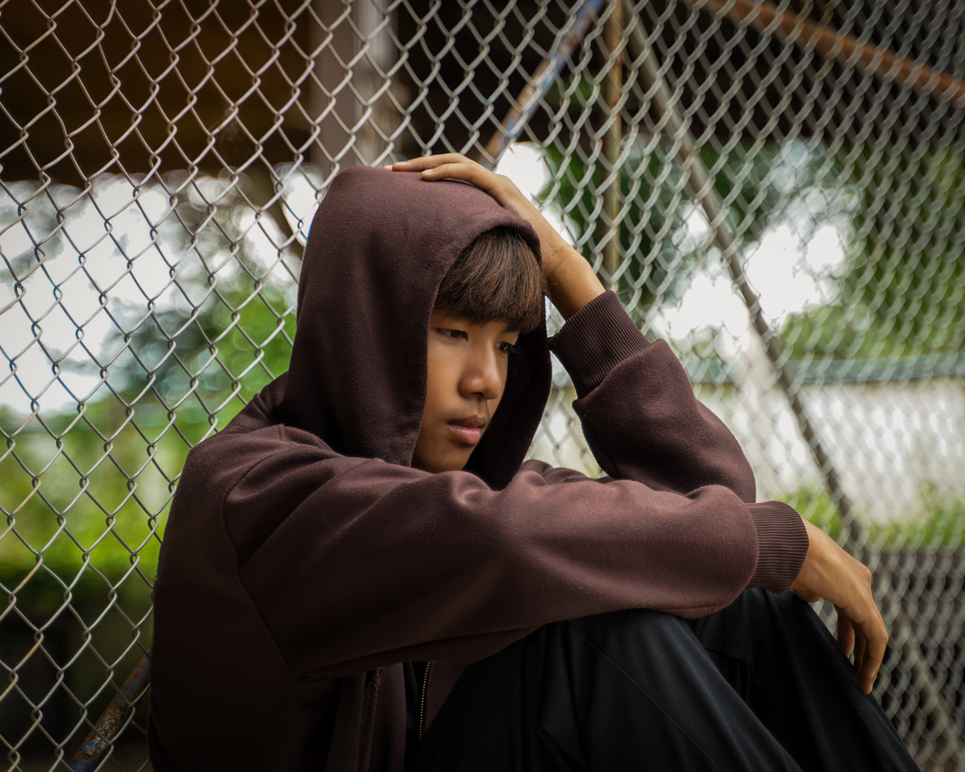 Asian boy wearing a hoodie sits with his knees bent, his back leaning against a wire mesh fence, his head in his hands. The concept of troubled teenagers, loneliness, and depression.