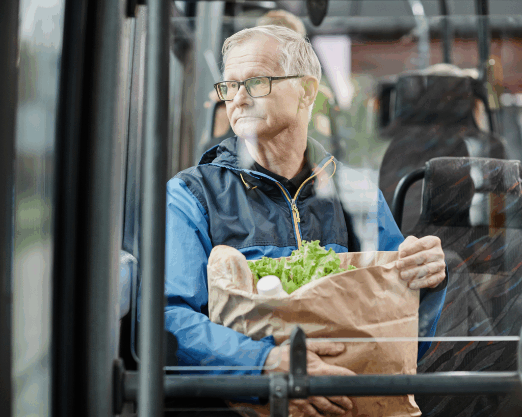 Senior man looking out a window on a bus while traveling by public transport in city and holding paper bag of food