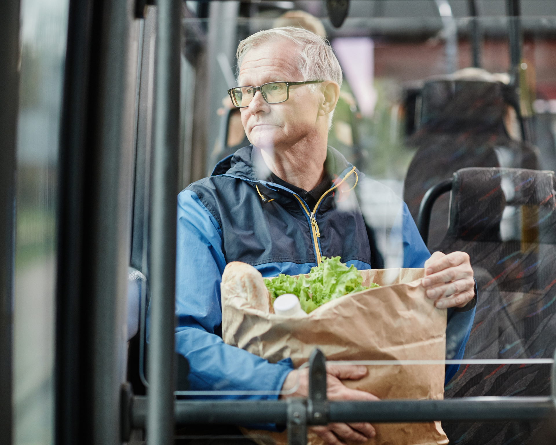 Senior man looking out a window on a bus while traveling by public transport in city and holding paper bag of food