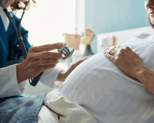Close up of a pregnant woman sitting during medical examination with female doctor holding stethoscope near belly, focusing on prenatal health care and pregnancy monitoring