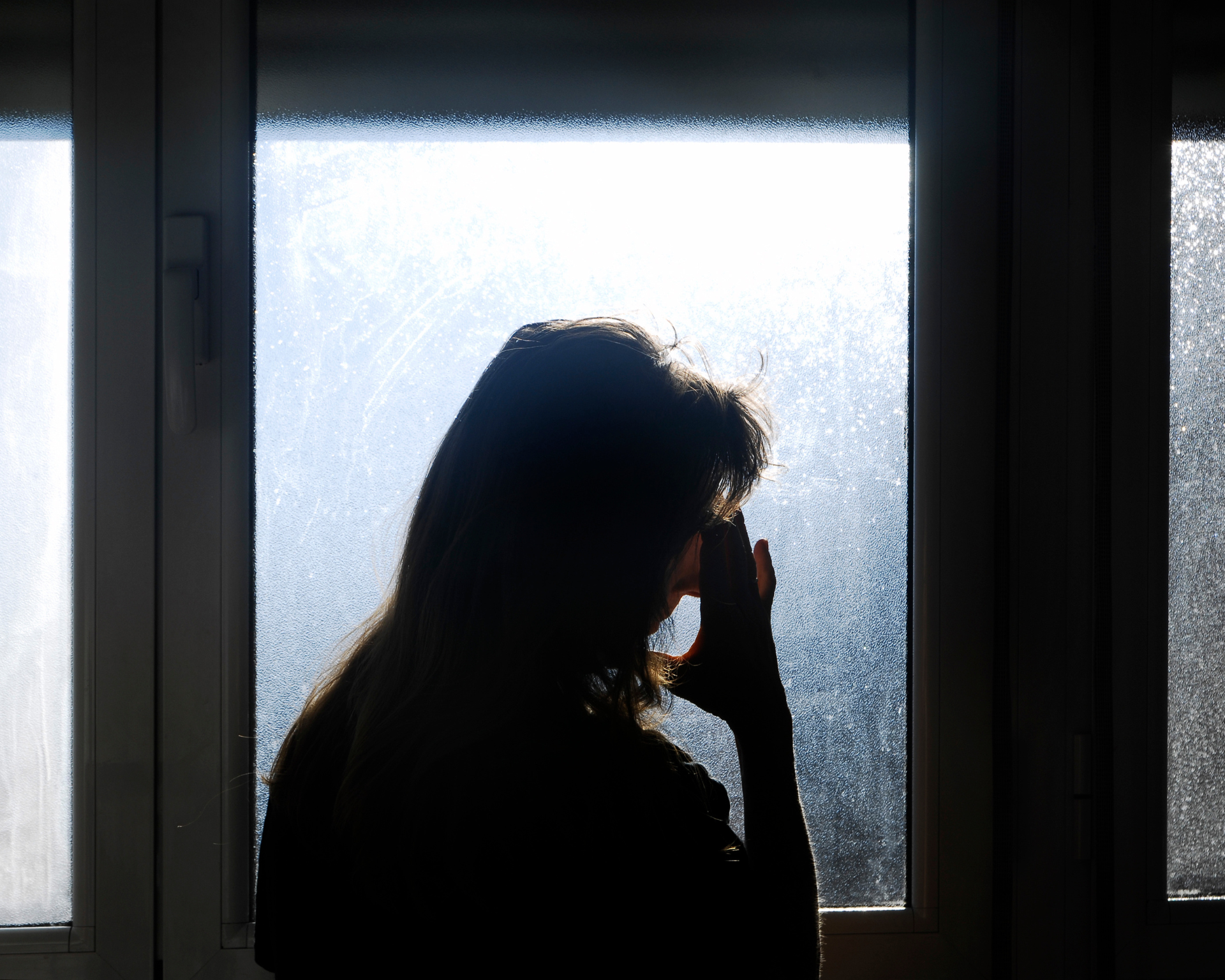 Silhouette of a woman standing in front of a translucent window with one hand placed on her forehead in a gesture of worry or pain