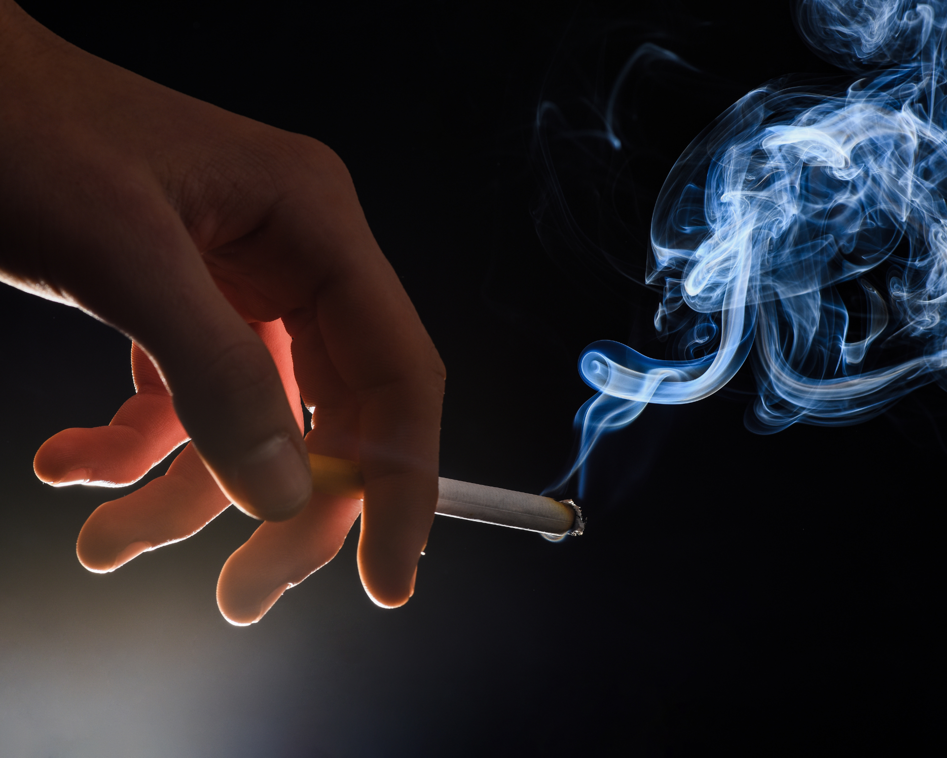 Close-up image of a male hand holding a burning cigarette with a swirl of smoke on a dark background. Tobacco concept