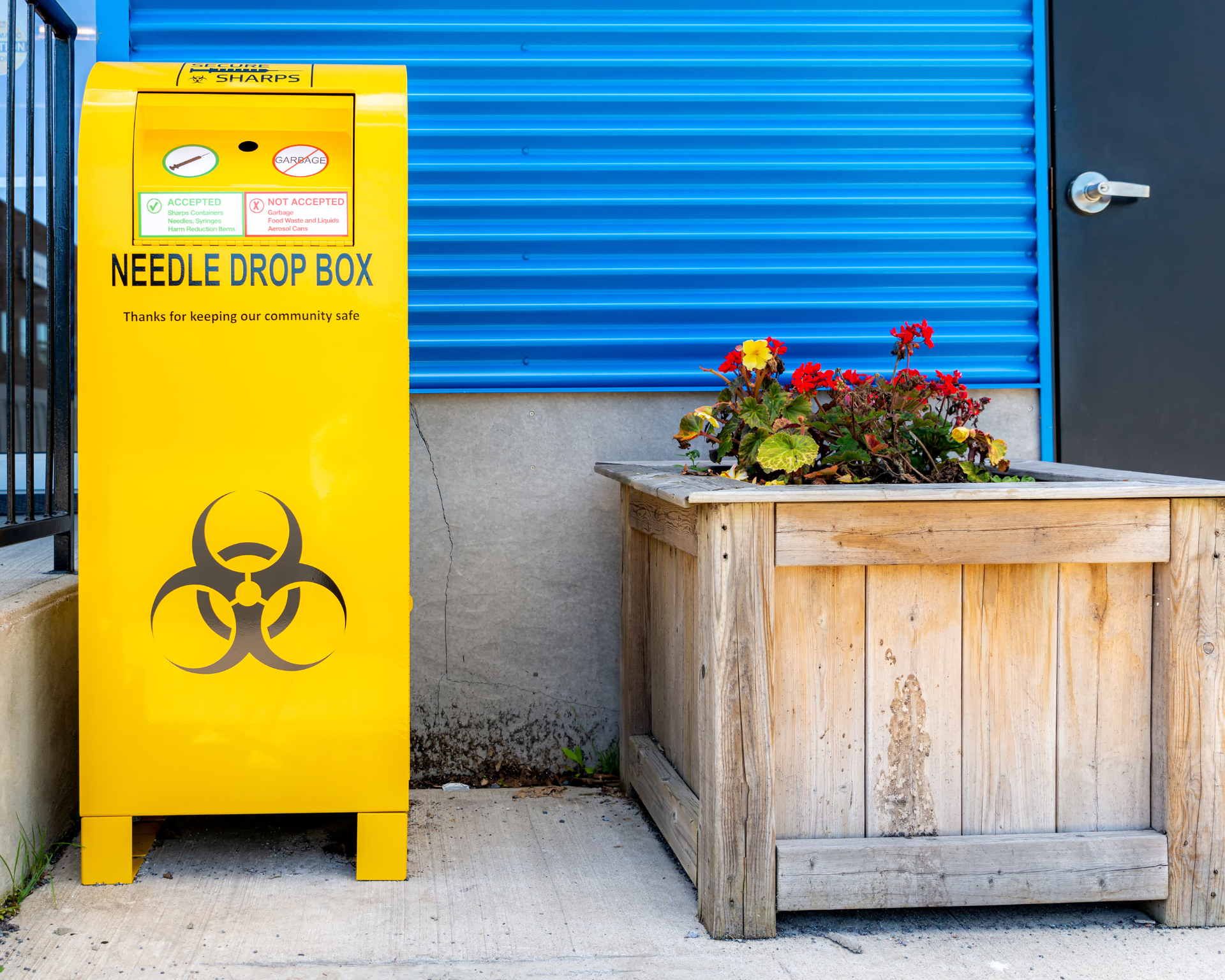 A public drop box for used needles, on a neighborhood sidewalk by a building. The box is bright yellow and bears the standard bio-hazard symbol.