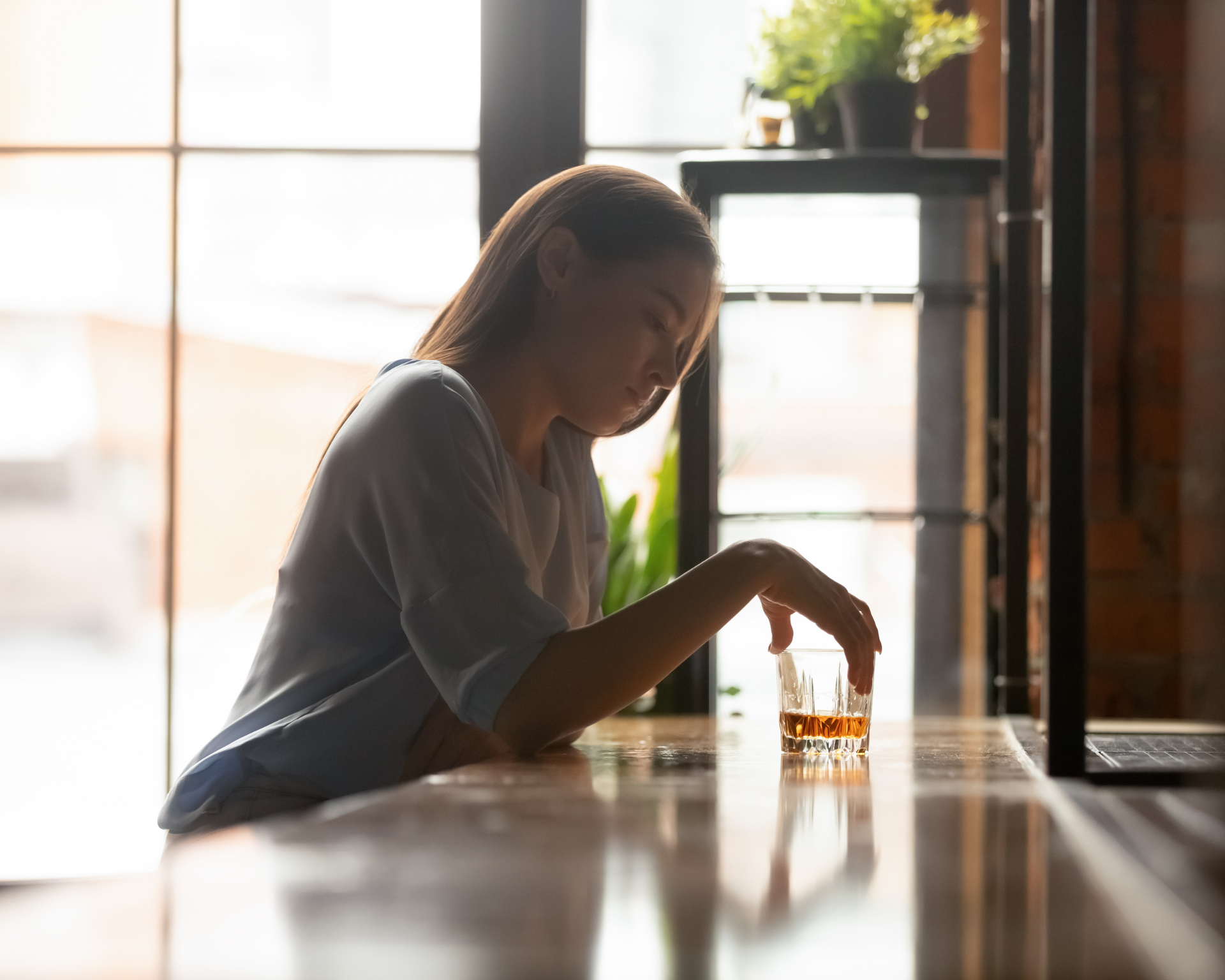 Young woman sitting on bar counter, looking down at glass full of alcohol
