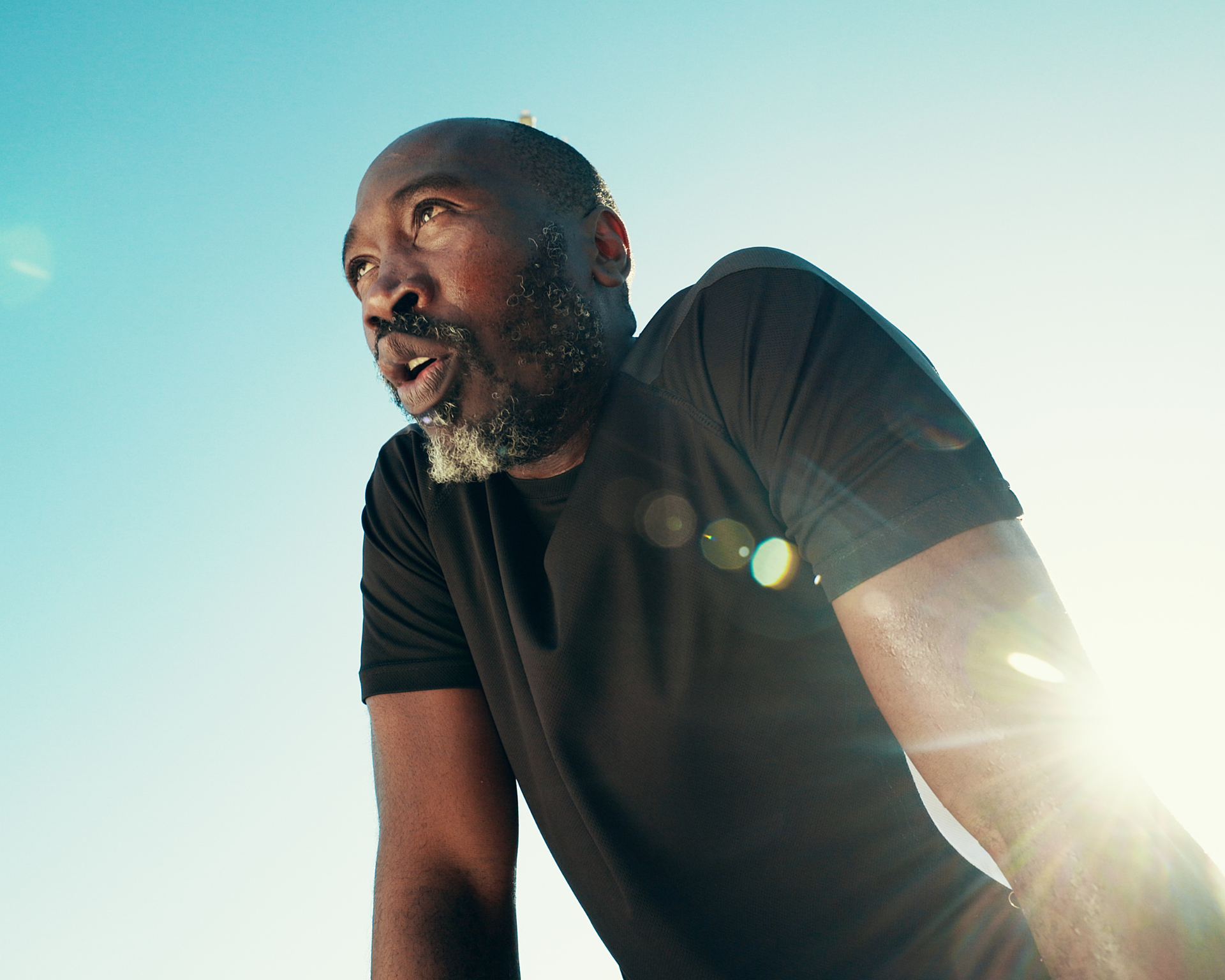 Breathing, fitness and black man by blue sky for running, workout or training for marathon with break. Fatigue, lens flare and African mature athlete with rest for outdoor cardio exercise from below.