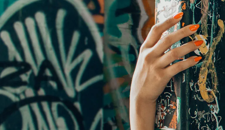 a hand with orange nail polish touching a wall with graffiti