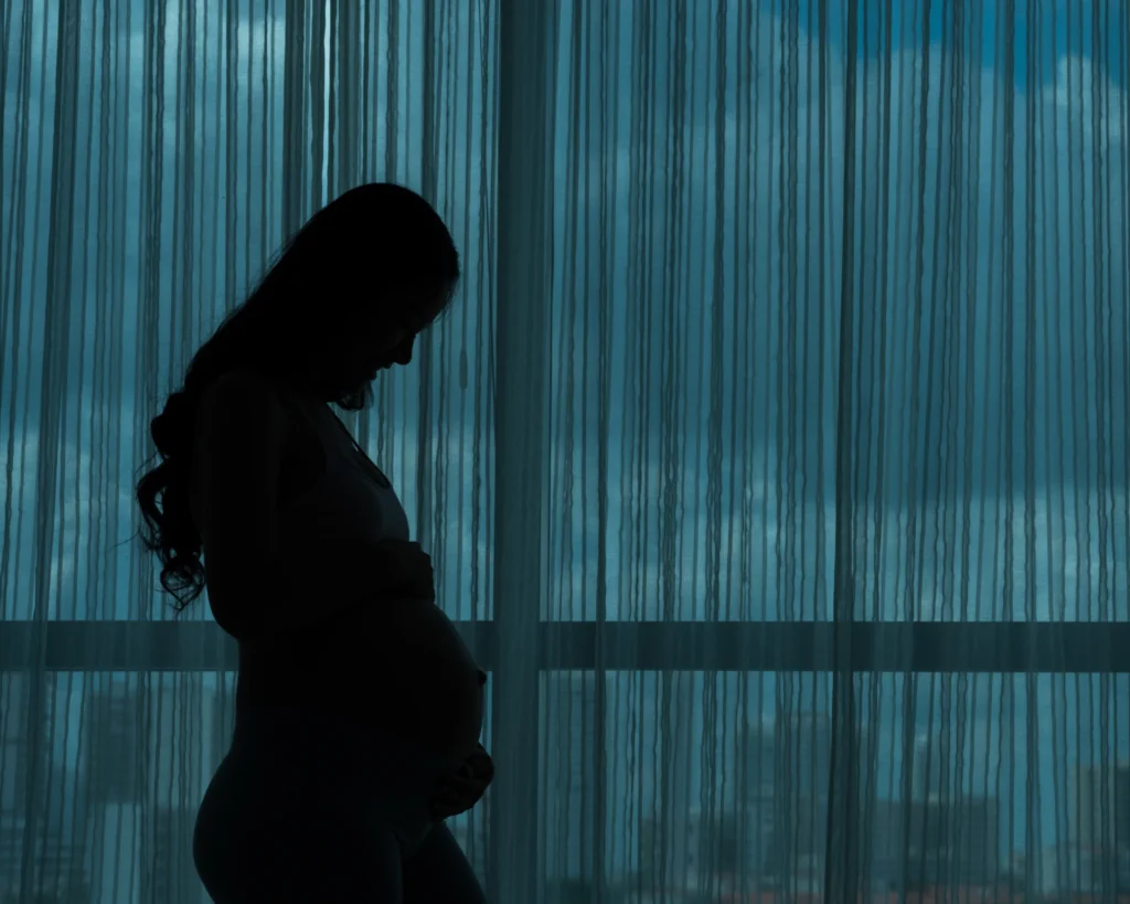 Silhouette of a young woman experiencing pregnancy, standing in front of a darkened window