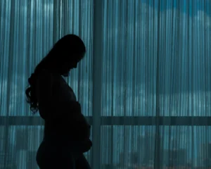 Silhouette of a young woman experiencing pregnancy, standing in front of a darkened window