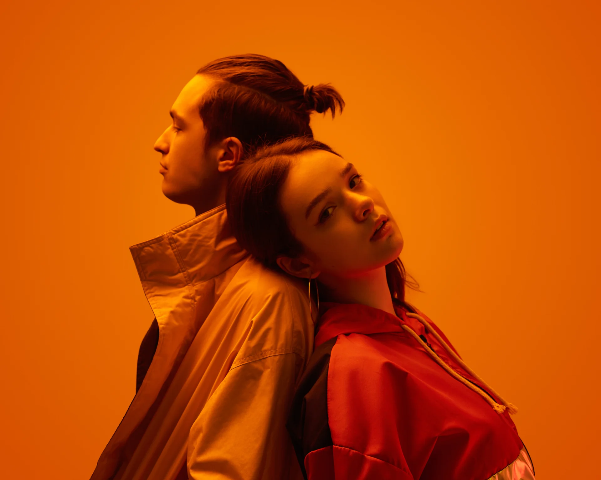 Portrait of the side view of a young couple in trendy clothes, standing back to back on an orange background in a studio with neon lights. Romantic couple concept