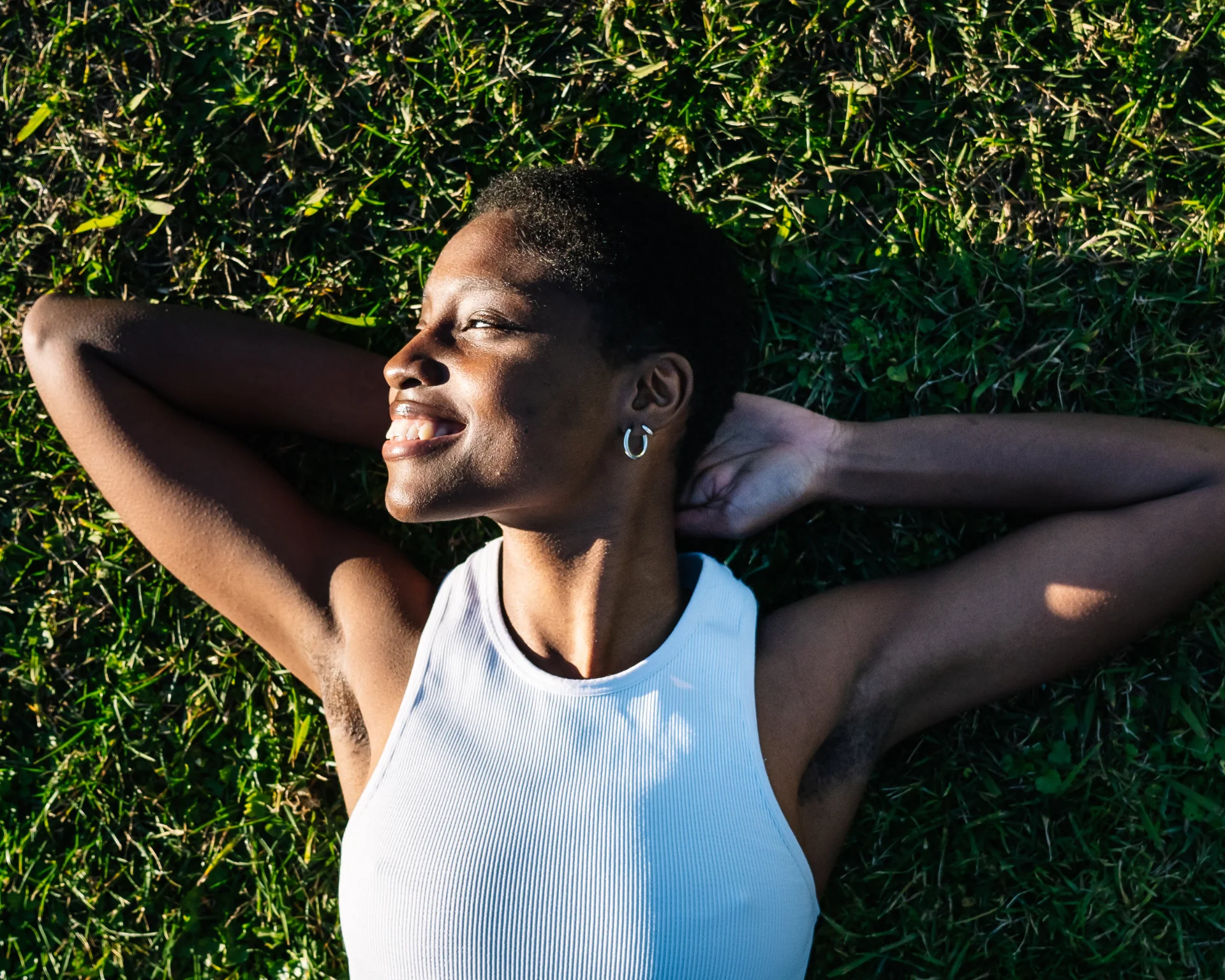 Serene young black woman enjoying a moment of peace lying on vibrant green grass, hands clasped behind her head, and a gentle smile on her face. Connecting with nature concept