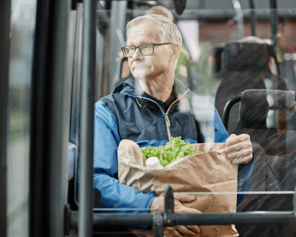 Senior man looking out a window on a bus while traveling by public transport in city and holding paper bag of food