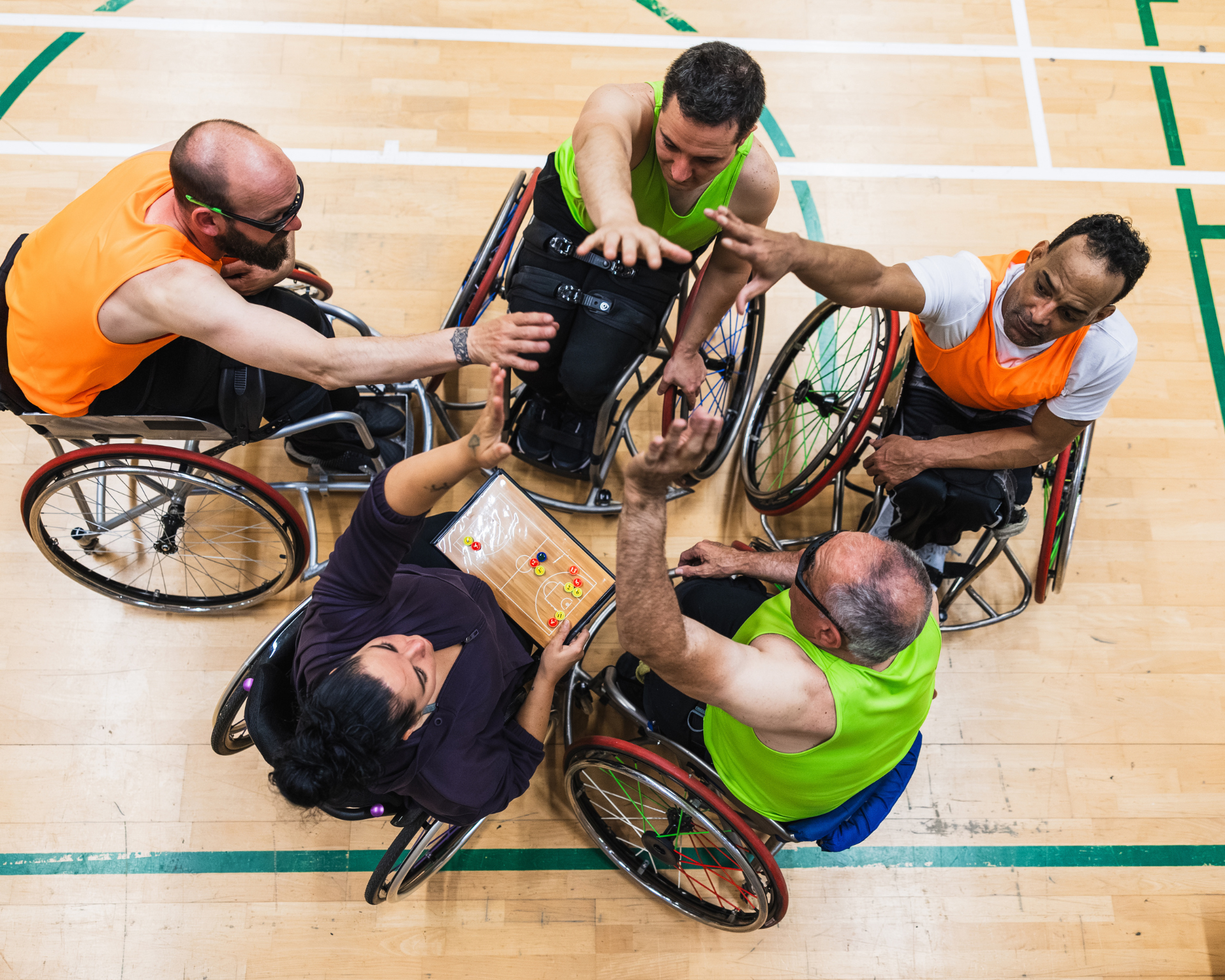 Five people in wheelchairs forming a huddle, reaching hands together, celebrating team spirit during a basketball game. One person is holding a tactics board. Concept for adults with disabilities participating in organized sports