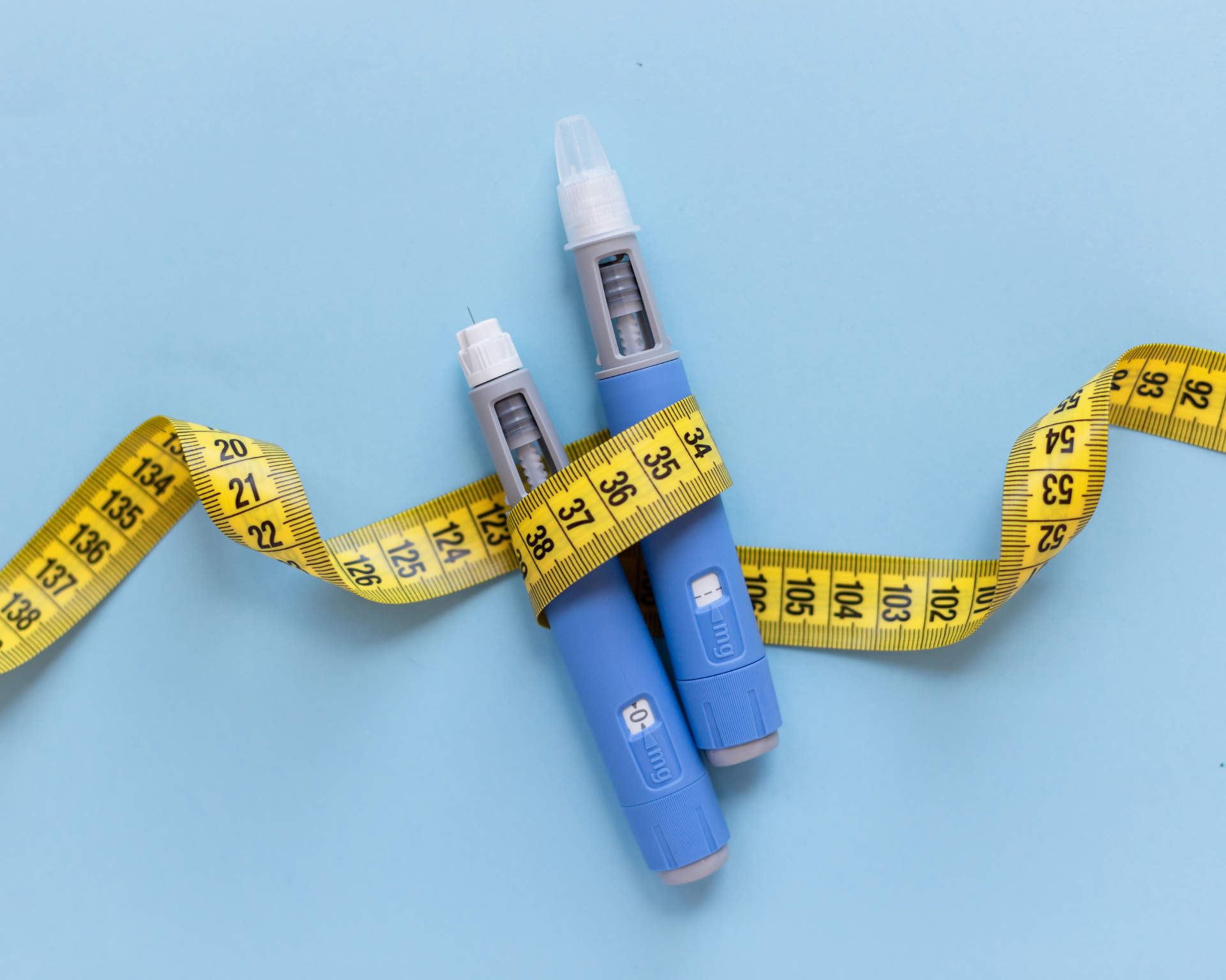 Two injectors dosing pens for subcutaneous injection of antidiabetic medication or anti-obesity medication hovering over a blue background. Yellow measuring tape around the injectors, suggesting weight loss.